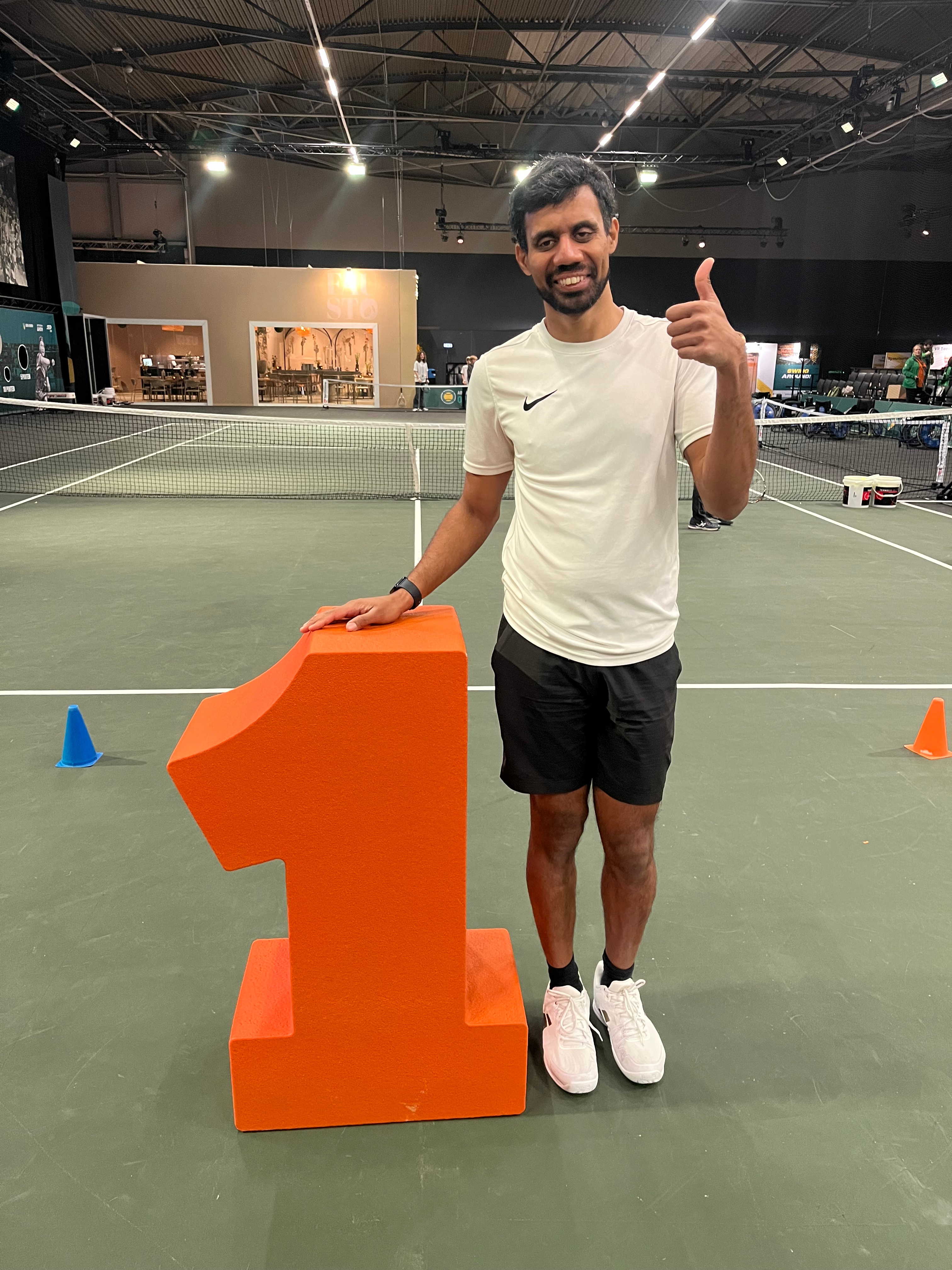 Naqi giving a thumbs up next to a large orange number one on a tennis court, with tennis nets and tennis equipment in the background.