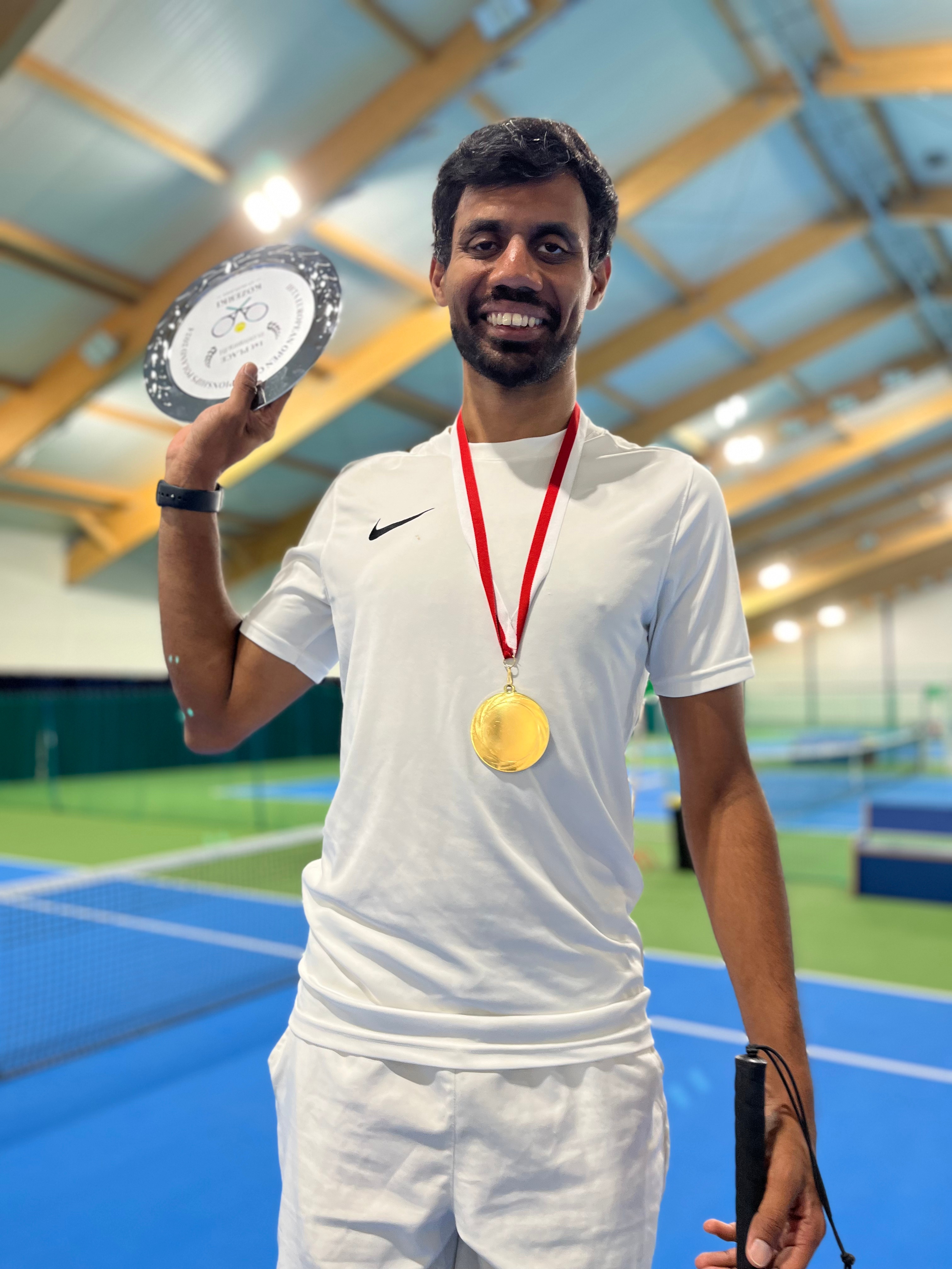 A smiling Naqi in a white athletic outfit holds a metal plate trophy in one hand and a gold medal on a red ribbon around his neck, standing on a tennis court with blue surfaces.